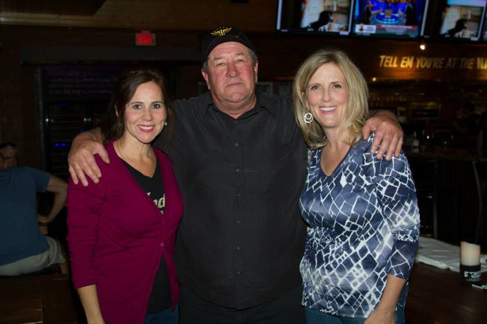 Three people pose together at a bar with televisions in the background; the two women smile while the man looks neutral.