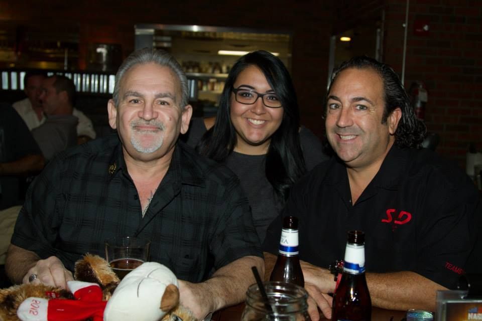 Three people smile in a dimly lit restaurant or bar, positioned behind a table with two beer bottles and a plush toy.