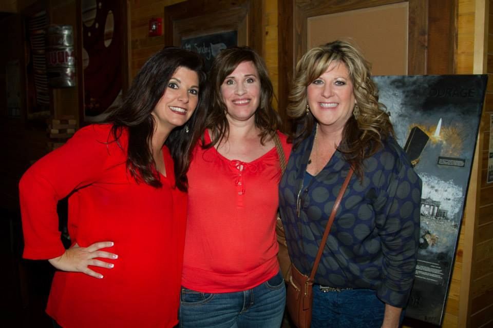 Three smiling people pose for a photo indoors, wearing casual tops in red and blue, standing against a wood-paneled wall.