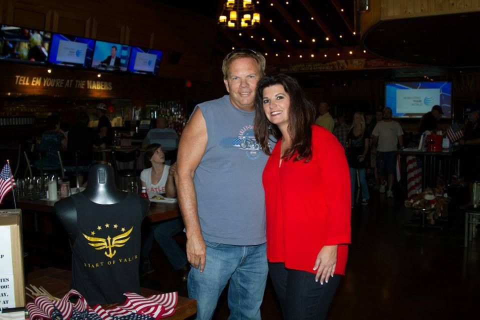 A man in a sleeveless shirt and a woman in a red top stand smiling in a restaurant bar with television screens above.