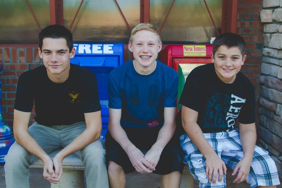 Three people sit side-by-side in front of blue and red newspaper stands against a brick wall.