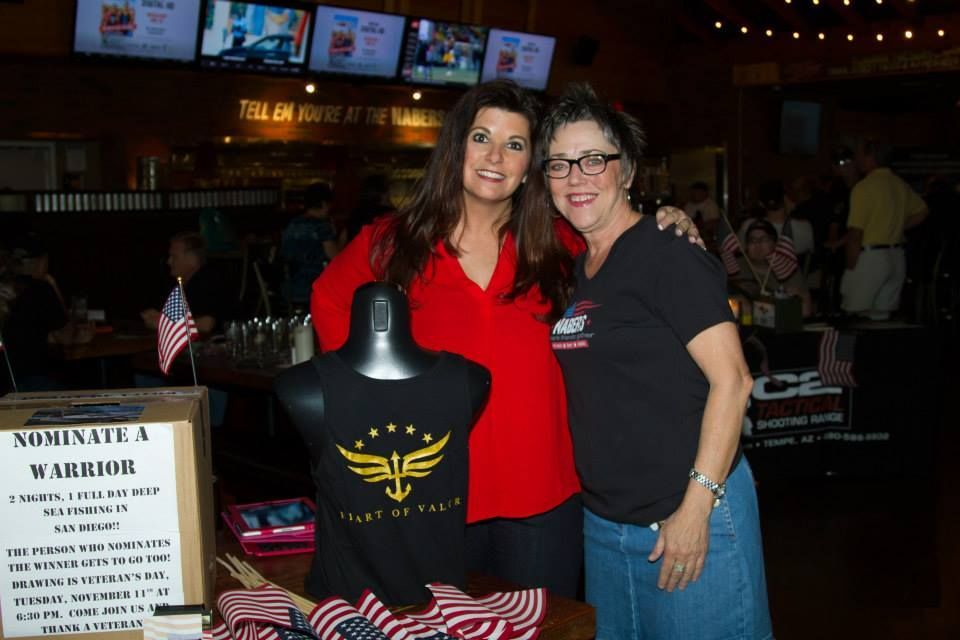 Two women smile while standing beside a display with a shirt and a “Nominate a Warrior” sign in a dimly lit bar setting.