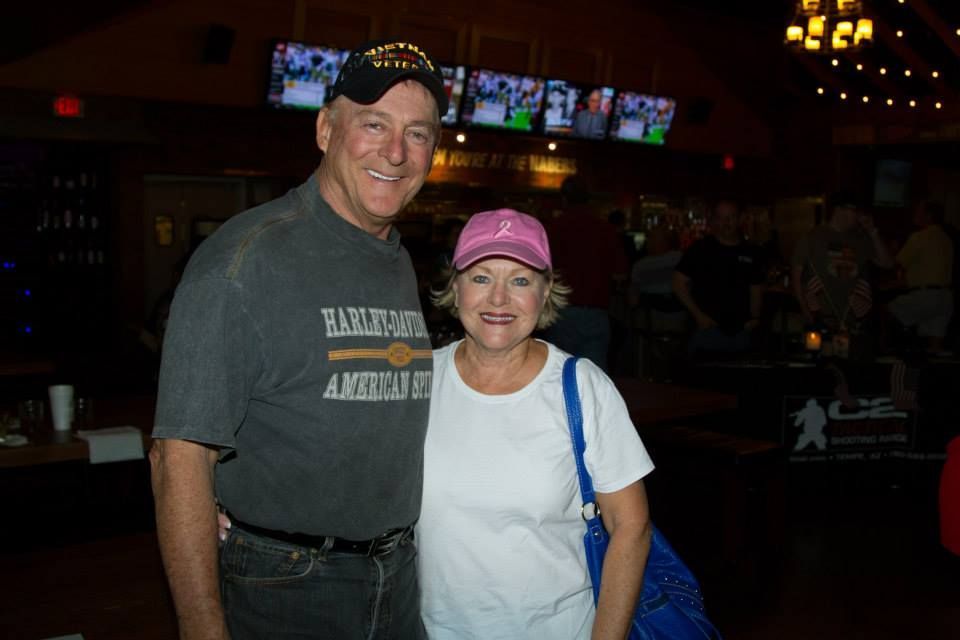 A man and woman smiling in a dimly lit venue, the man wearing a Harley-Davidson t-shirt and hat, the woman a pink cap.