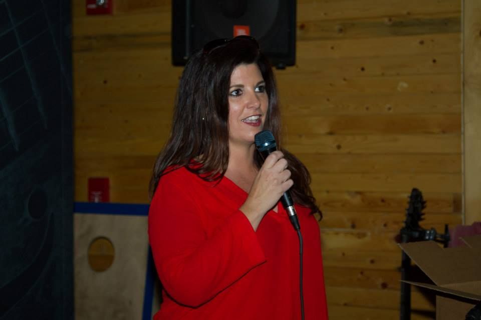 A person in a red top holding a microphone and speaking in front of a wooden wall.