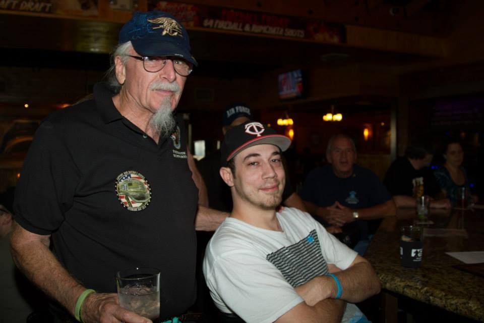 Two men standing together at a bar, one holding a glass and the other with arms crossed, in a dimly lit setting.