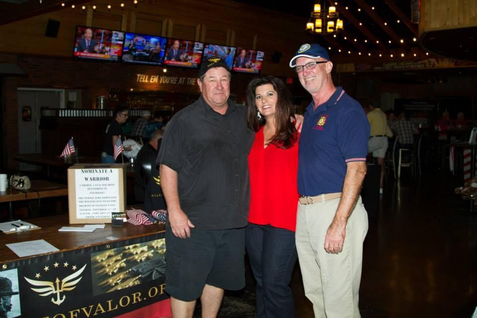 Three people stand together in a dimly lit venue, smiling for a photo in front of a table with a sign for HoeOfValor.org.