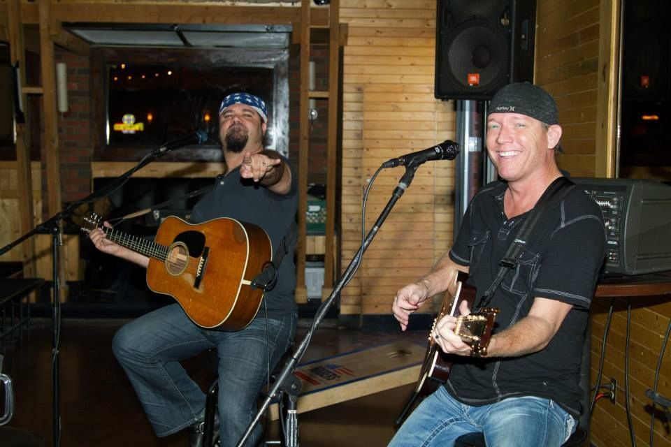 Two musicians play acoustic guitars and smile while performing on stage in a wood-paneled room.