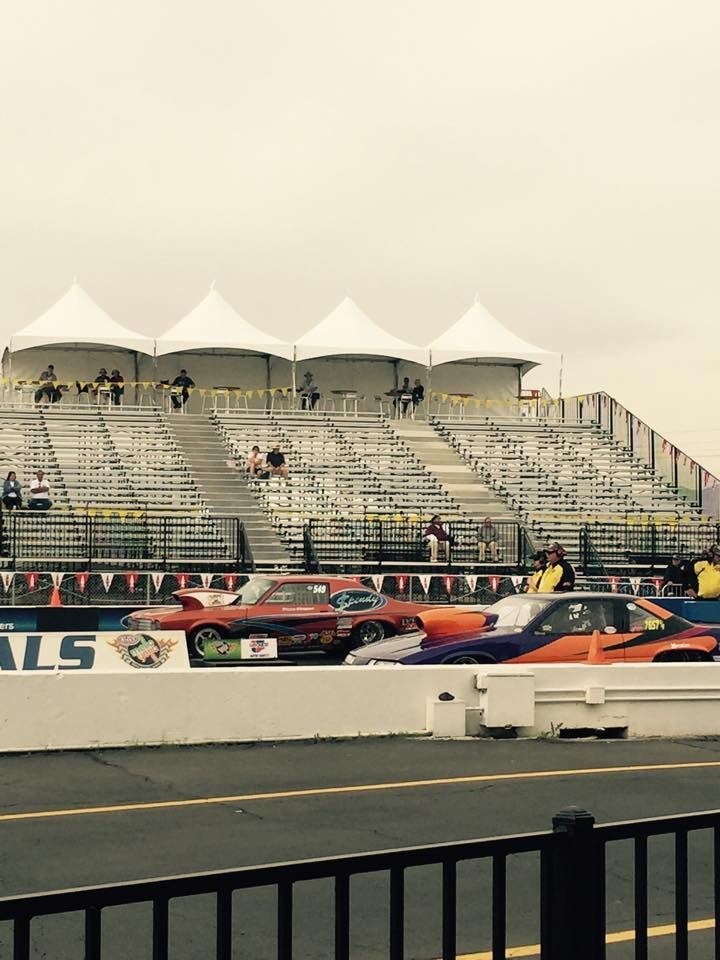 Two race cars driving side-by-side on a track in front of grandstand seating under white tent canopies.