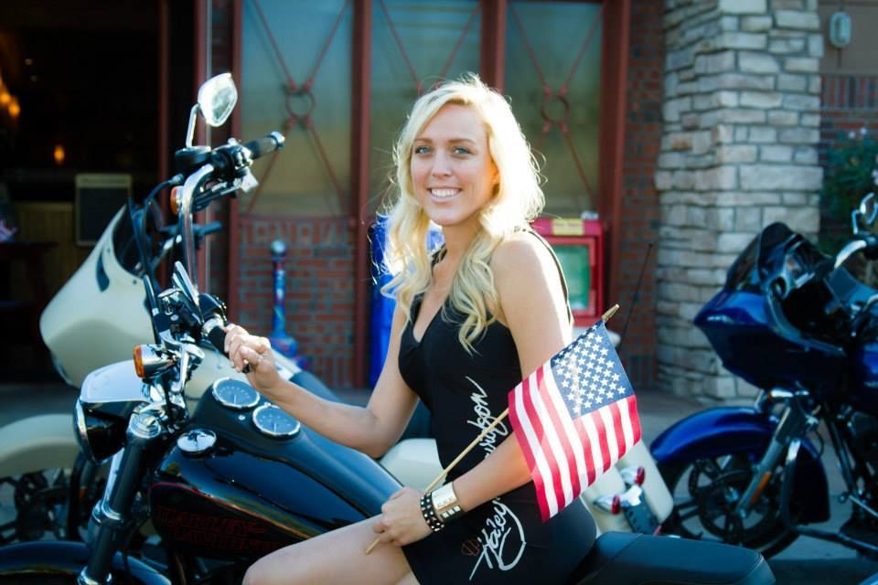 A blonde woman smiles while sitting on a black motorcycle, holding a small American flag in front of a brick building.