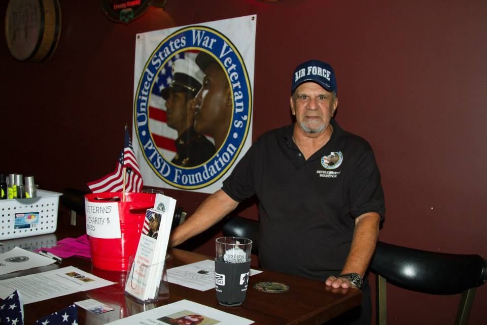 A man wearing an Air Force cap stands behind a table with materials for the United States War Veteran's PTSD Foundation.