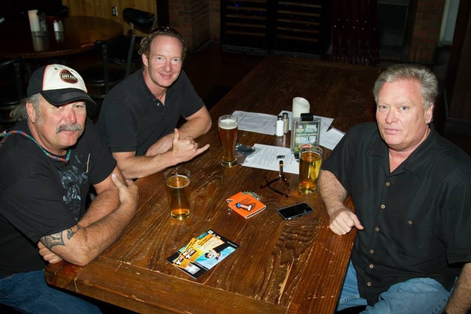 Three men sit at a wooden restaurant table with glasses of beer, salt shakers, and a brochure.