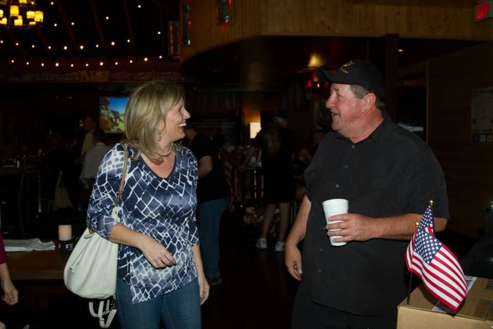 A woman and a man in a hat talking and laughing in a dimly lit, rustic bar setting with a small American flag nearby.