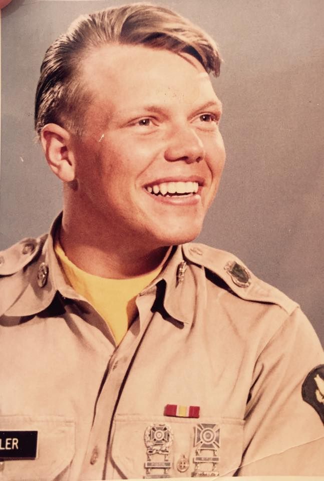 A person wearing a tan military-style uniform with patches and a ribbon, smiling against a plain backdrop.