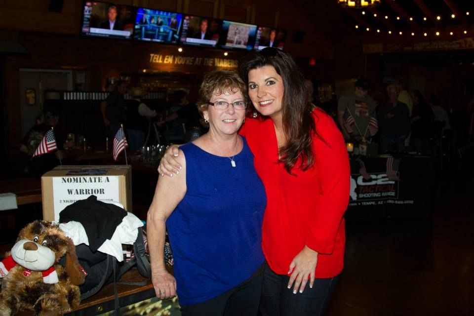 Two smiling people stand together in a restaurant with televisions and an American flag-themed display in the background.