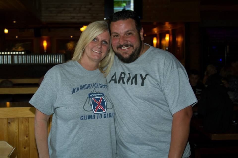 A smiling couple wearing grey t-shirts with Army insignia posing for a photo in a dimly lit indoor setting.