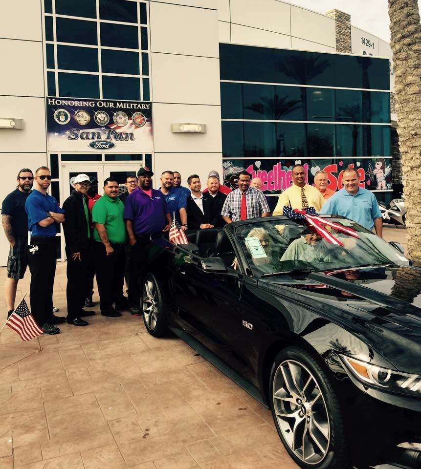 A group of people standing outside a San Tan Ford dealership next to a black convertible Mustang decorated with flags.