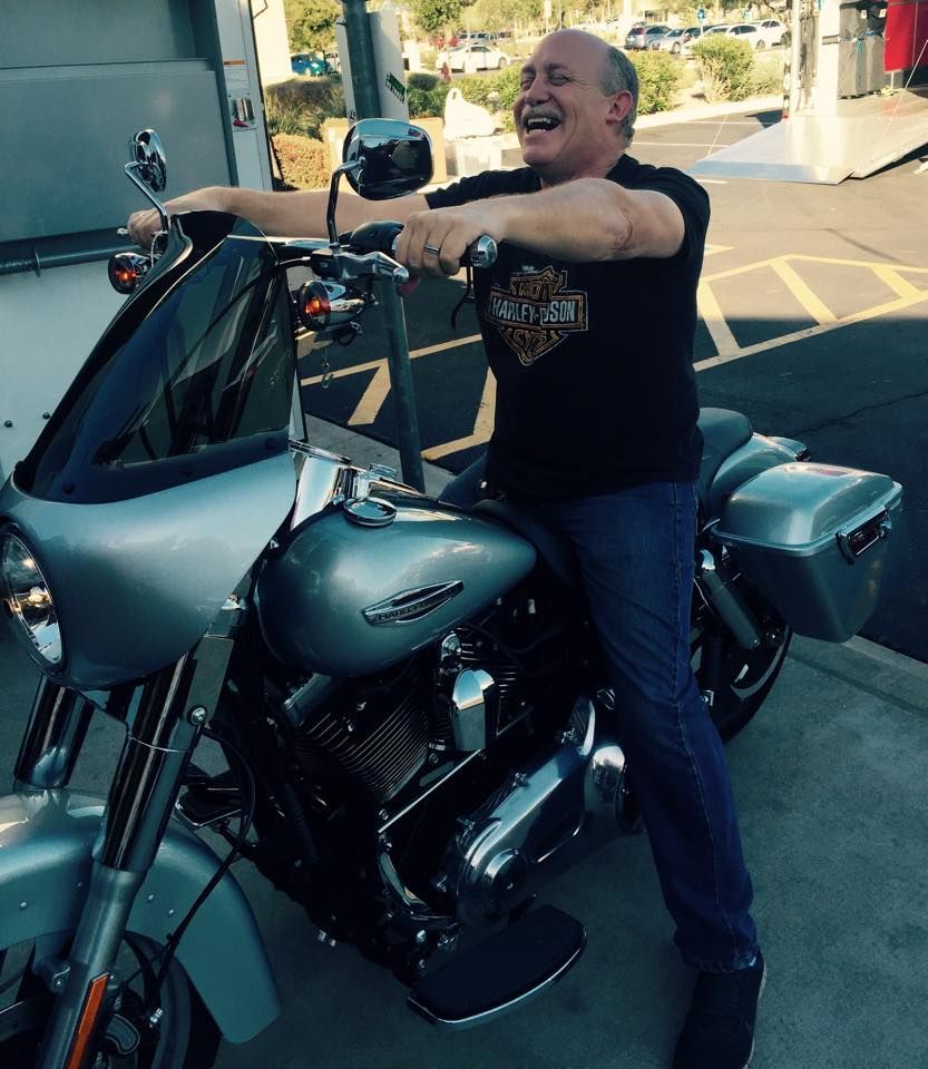 A smiling person wearing a black t-shirt sits on a silver Harley-Davidson motorcycle in a parking lot.