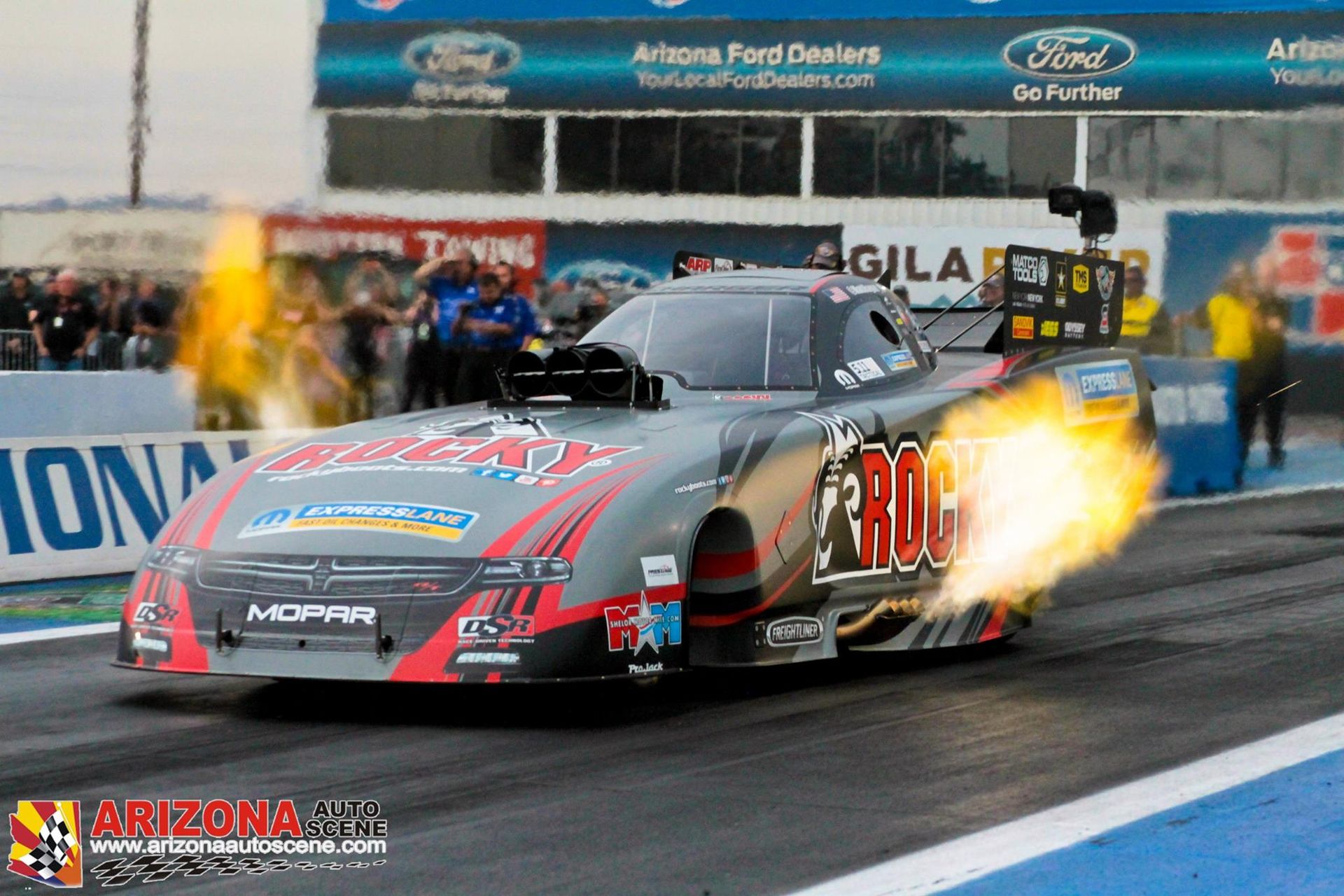 A gray Top Fuel dragster speeds down a race track with bright orange flames shooting from its side exhaust.