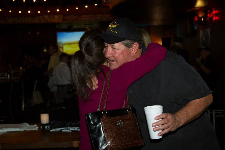 A person in a magenta top hugs a man in a black shirt and cap who is holding a white cup in a dim, social setting.