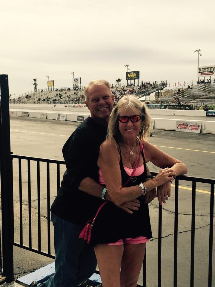 A couple embraces while standing behind a black metal fence at a sunny outdoor racetrack with stadium seating.
