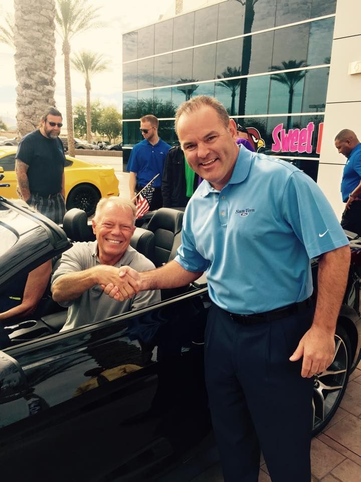 Two men shake hands beside a convertible car in a parking lot, with onlookers in the background.