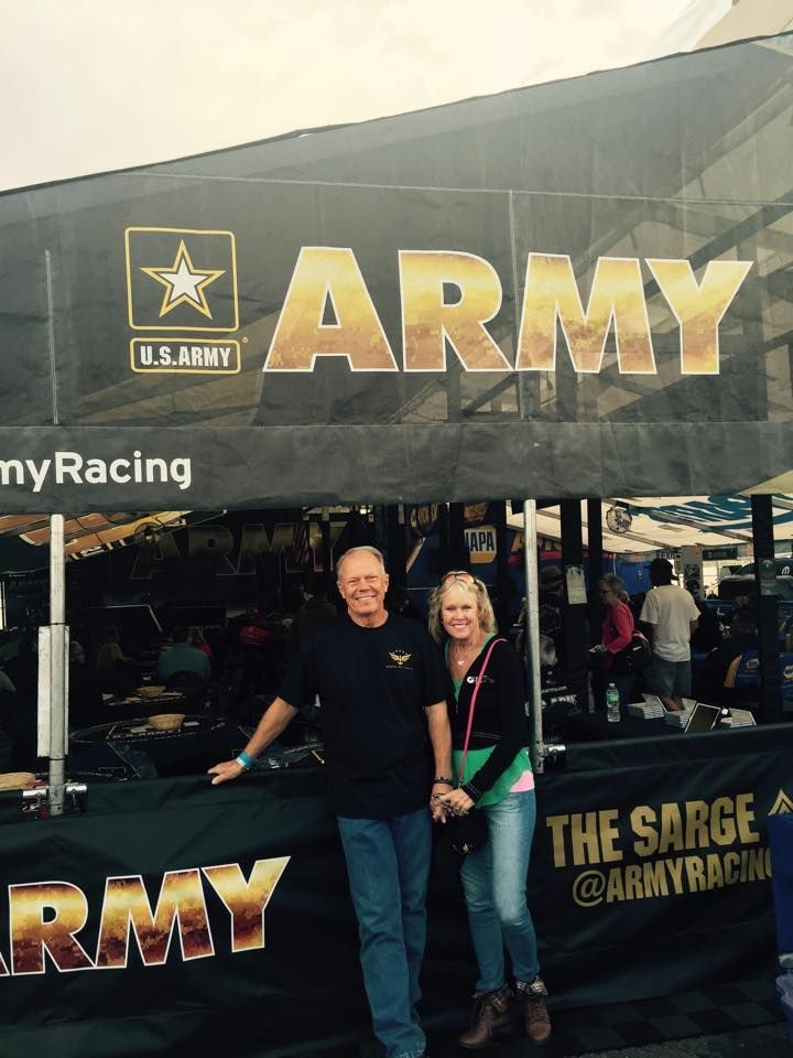 A smiling pair stands in front of a black U.S. Army Racing tent at a public event.