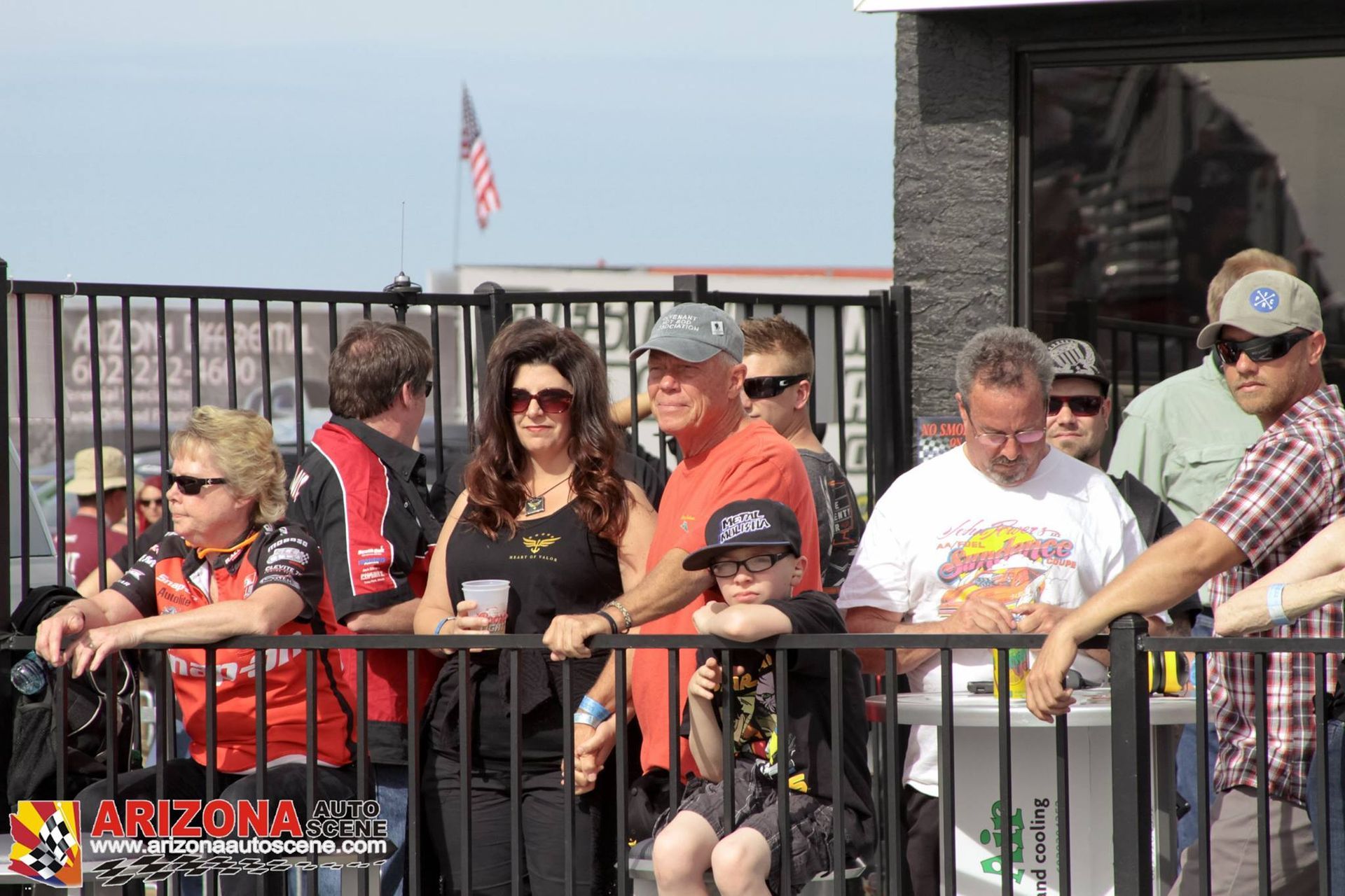 A group of people watch a race from behind a black fence, with an American flag flying in the background.