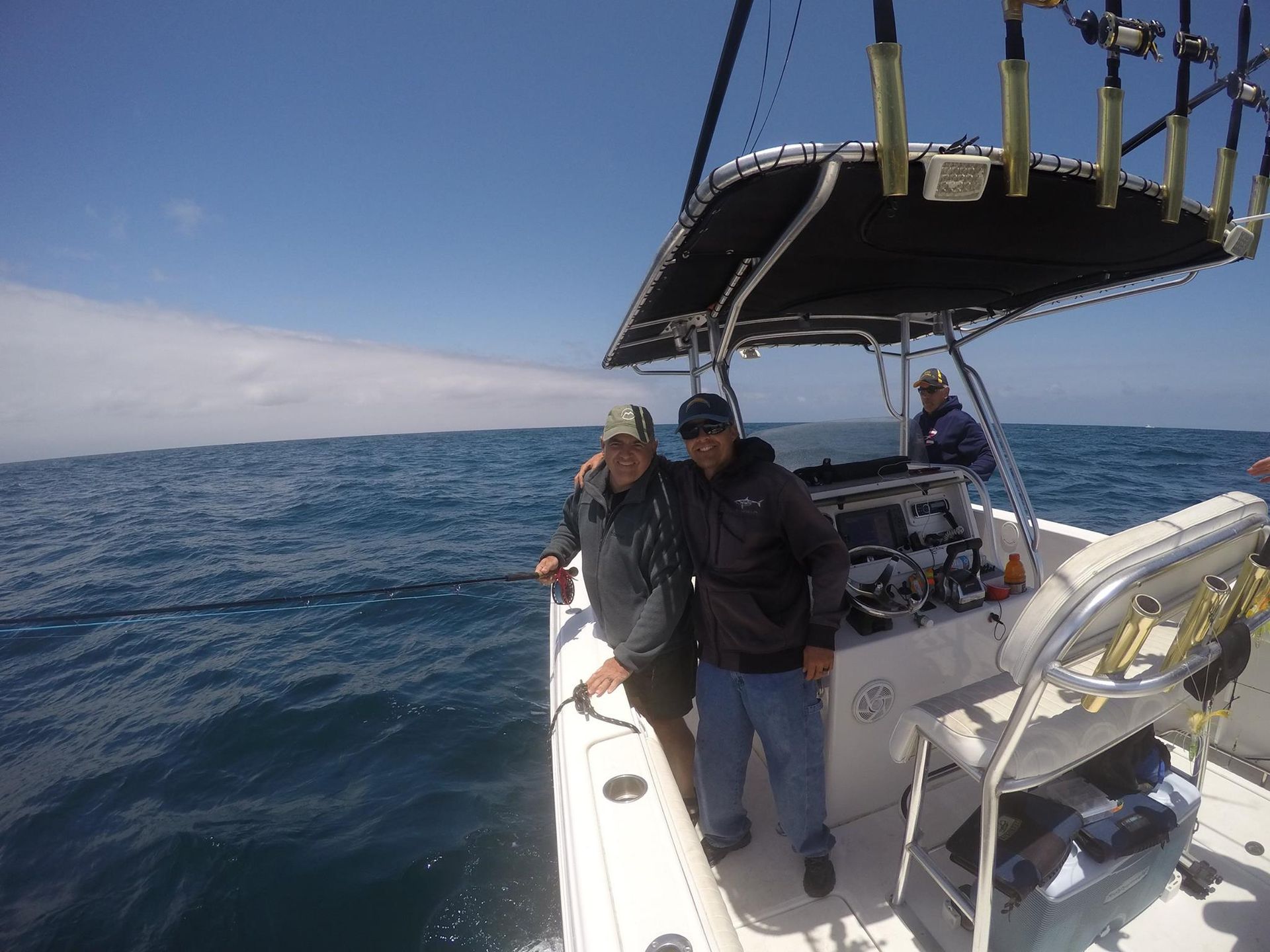 Two men on a boat, posing for a photo, with ocean backdrop. One is holding a fishing rod.