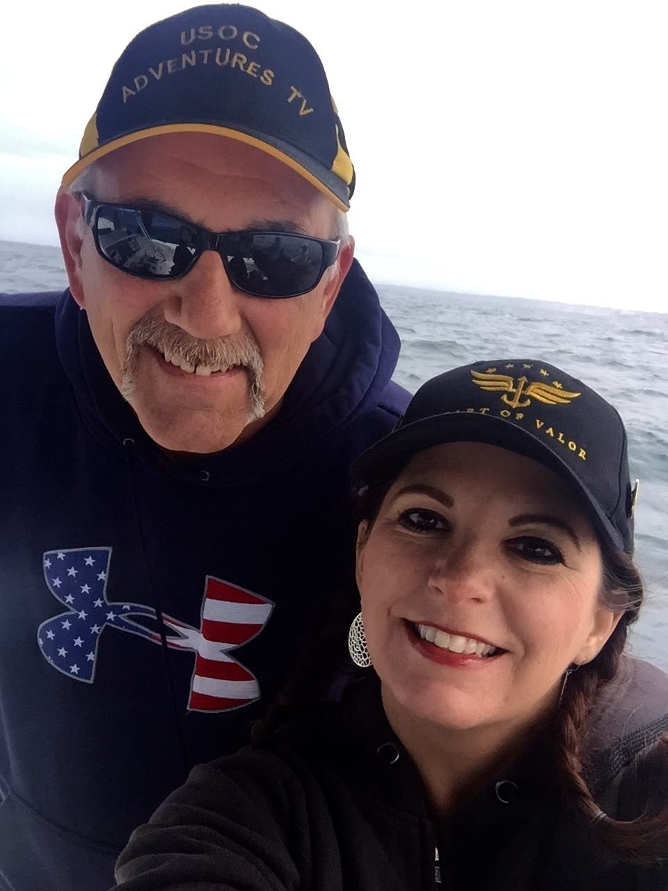 Man and woman on a boat, smiling. Man in sunglasses, cap. Woman with cap, dark hair. Water in the background.