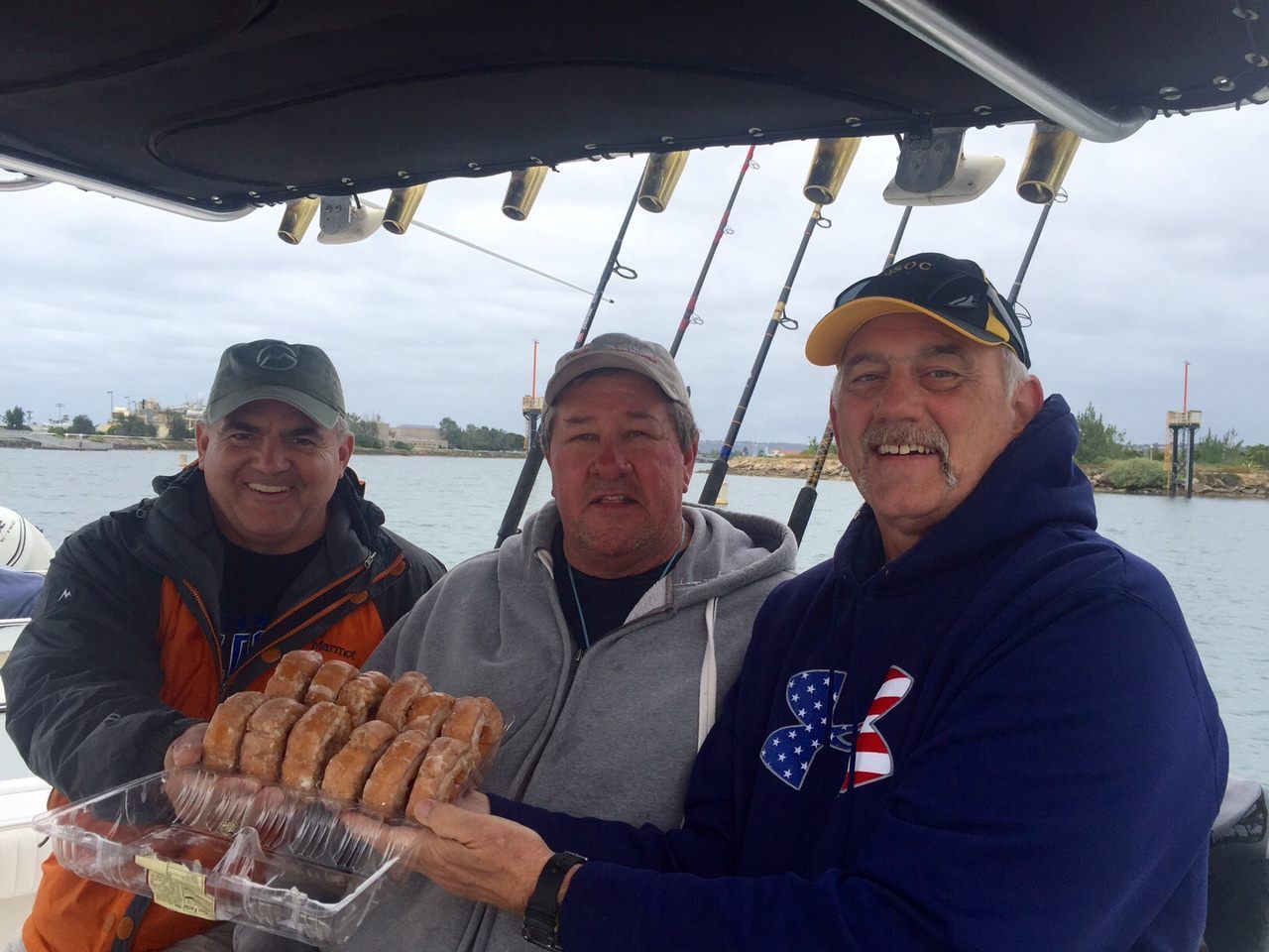 Three men on a boat holding donuts, smiling. Fishing rods visible, overcast sky.