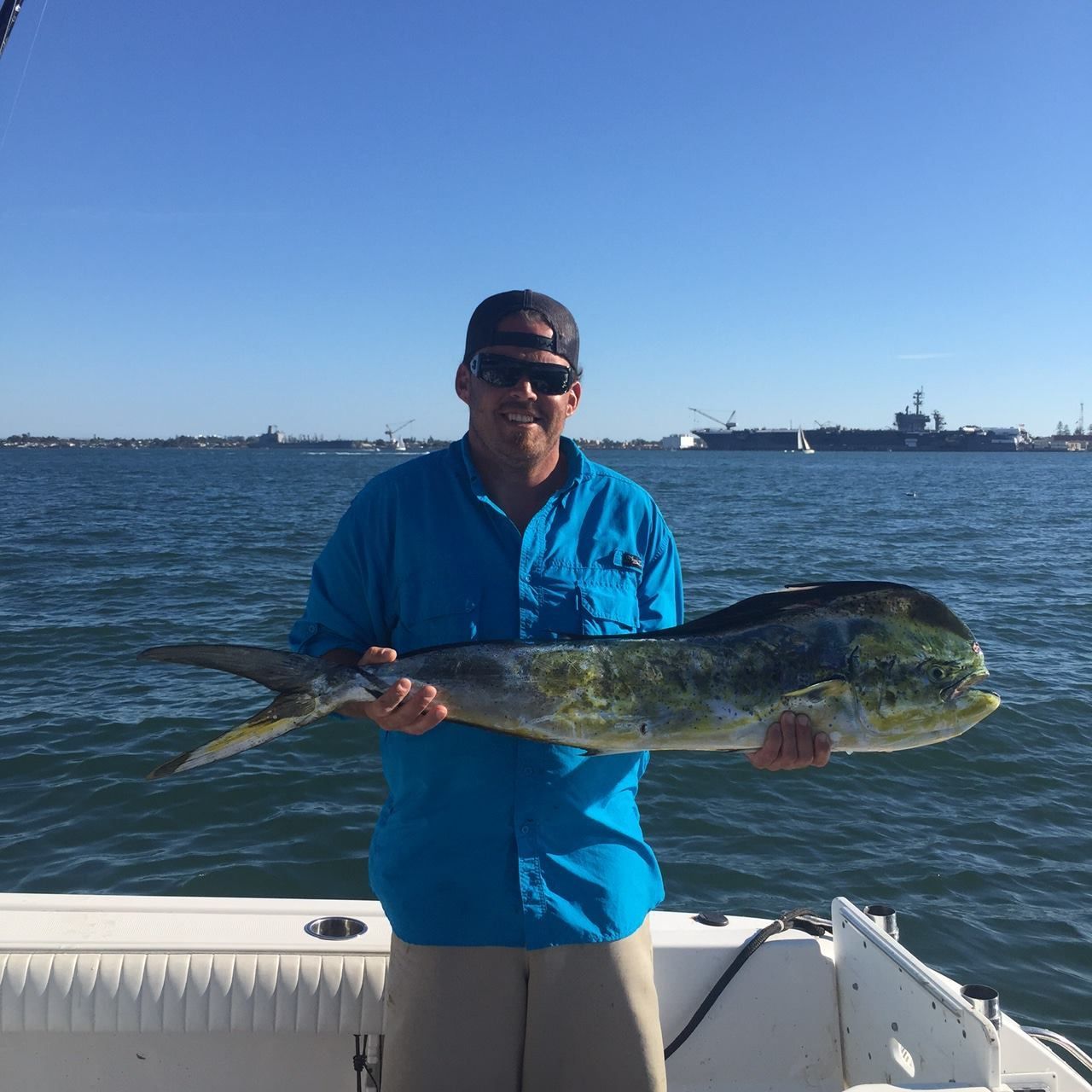 A person in a blue shirt and sunglasses holds a large mahi-mahi fish on a boat, with a navy ship in the background.
