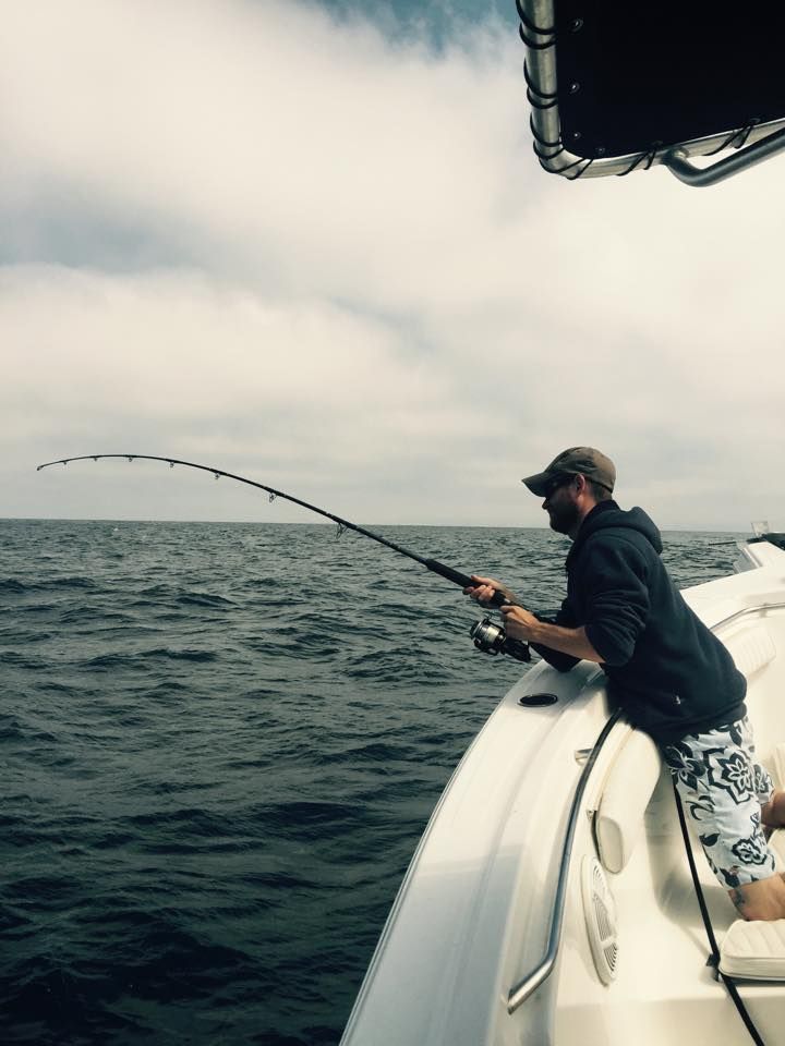 A person in a dark hooded sweatshirt and patterned shorts stands on a boat, holding a fishing rod bent with a catch.