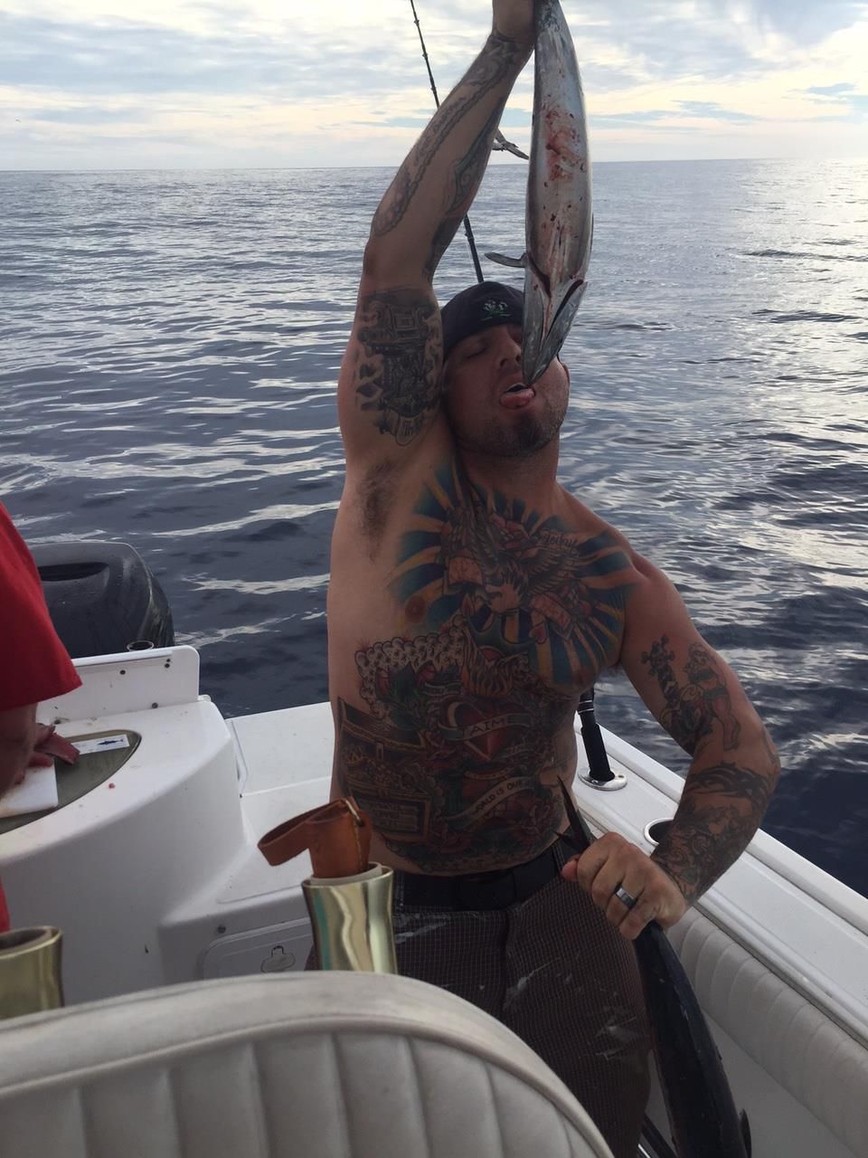 A shirtless tattooed person holds a fish up to their mouth while on a boat in the ocean.
