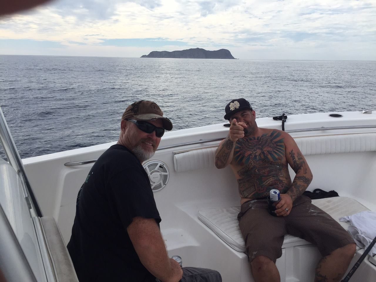 Two men on a boat, pointing at camera, island in the background, cloudy sky.