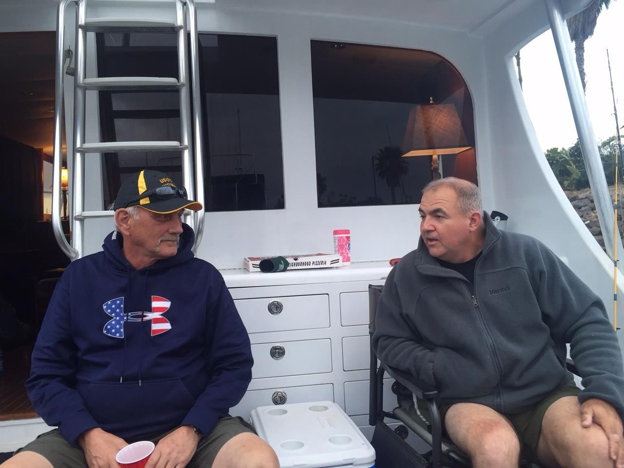 Two men on a boat deck, talking. One in a navy sweatshirt with an American flag, the other in a gray jacket. White storage behind them.