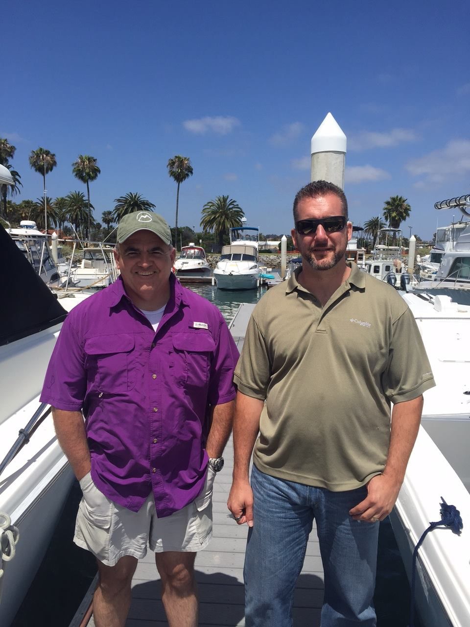 Two men on a dock in front of boats on a sunny day. One wears purple, the other green.