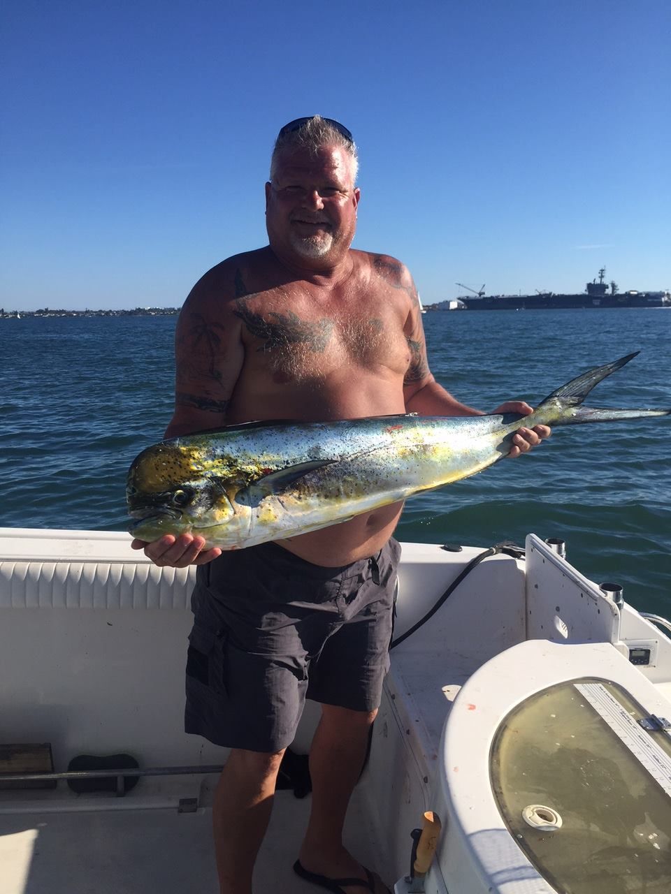 A person holds a large, brightly colored mahi-mahi fish on a boat, with a ship visible in the background on the water.