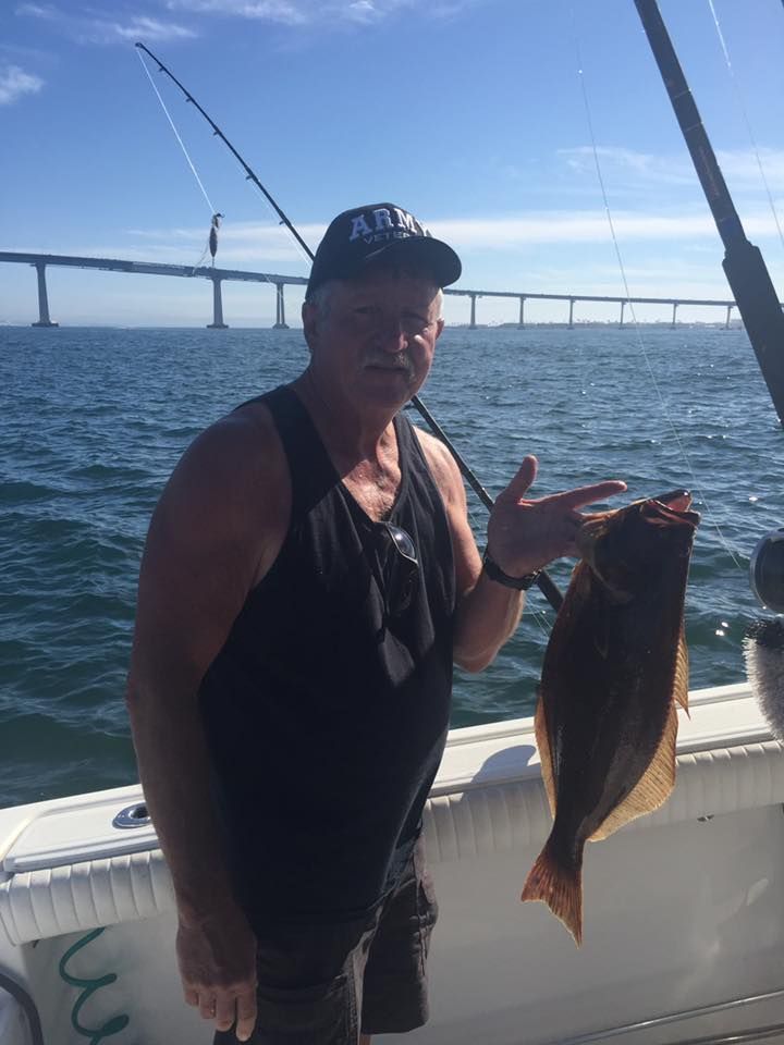 Man on boat holding fish, pointing, with a bridge in the background. Sunny day, water, fishing rods.
