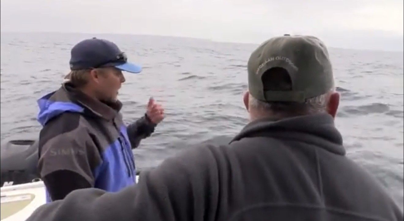 Two men on a boat at sea; one points towards the water, cloudy sky, cold weather.