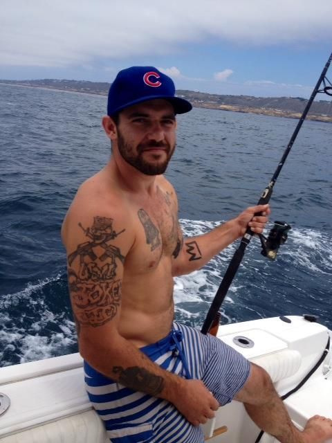 A man with arm tattoos wearing a blue baseball cap and striped shorts fishes from a boat on the open ocean.