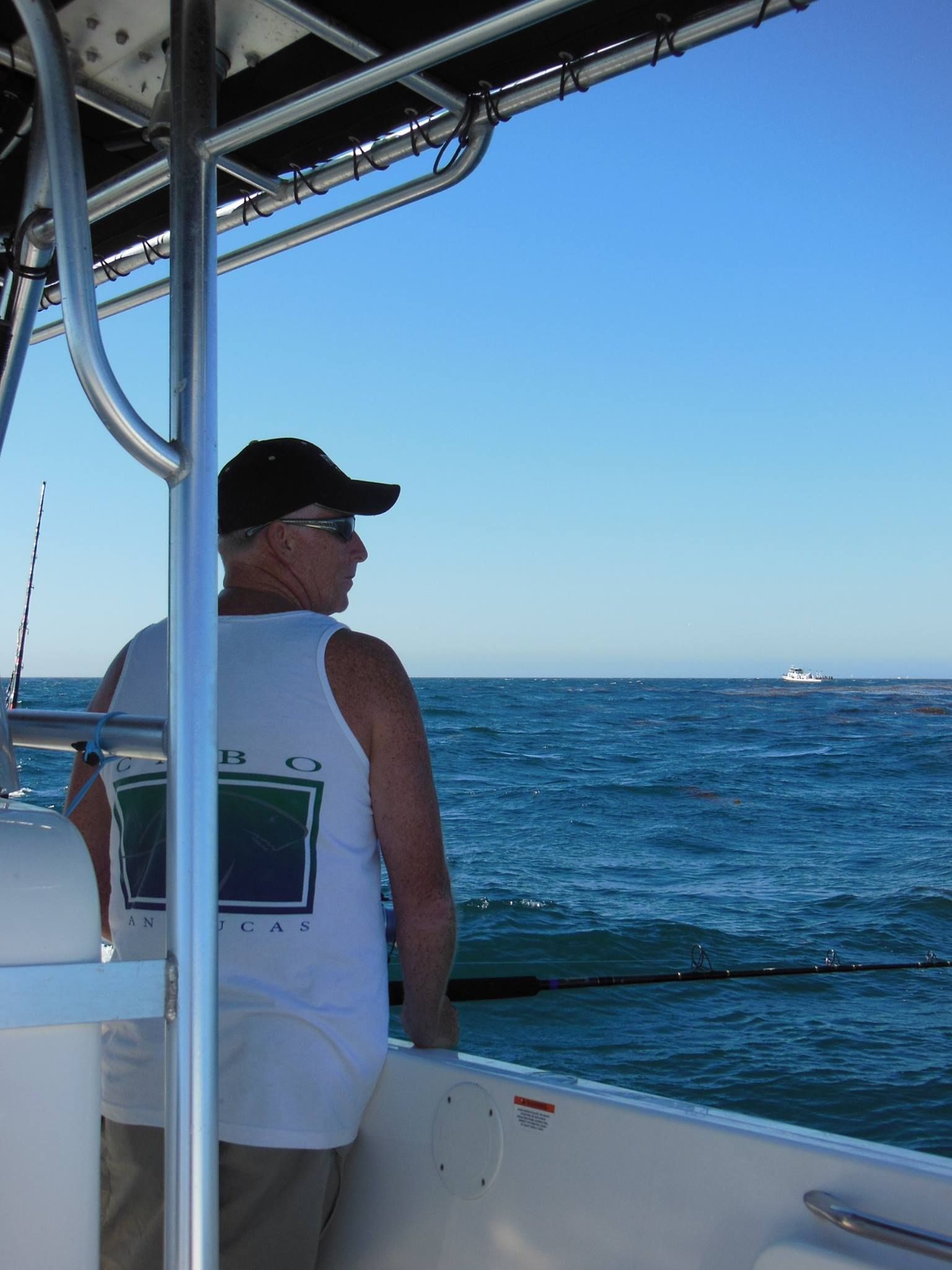 A person wearing a white tank top and black cap fishing from a boat on the open ocean under a clear blue sky.
