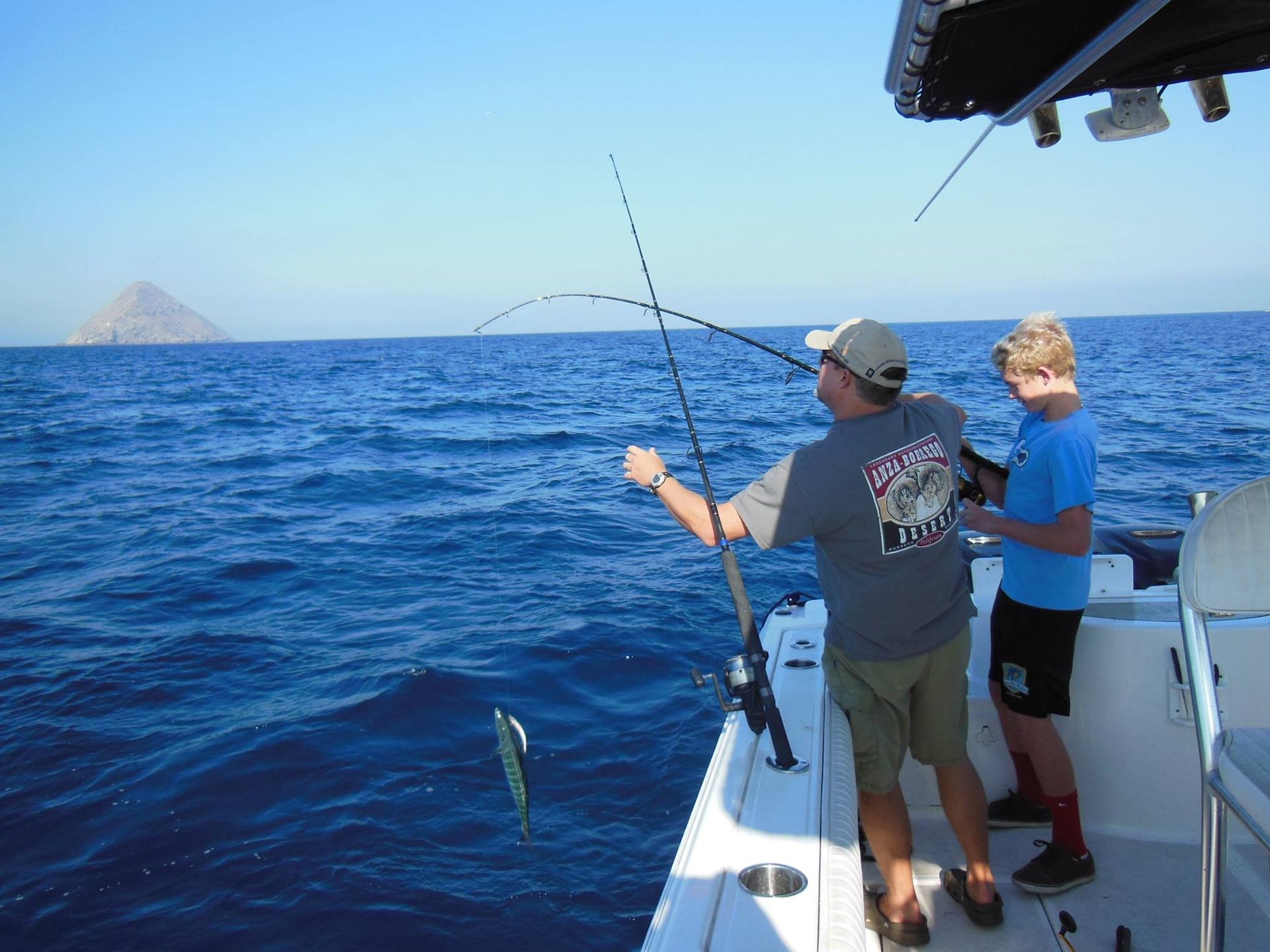 Two people stand on a boat, reeling in a fish from the open blue ocean near a distant rock formation.