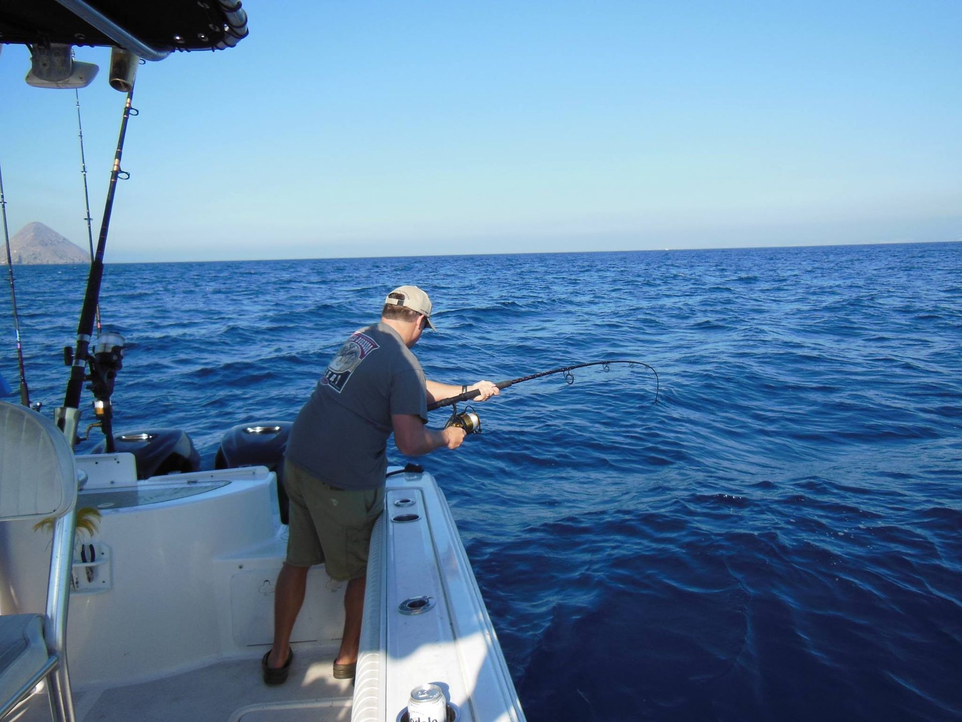 Person fishing from a boat on a sunny day. He holds a fishing rod over blue ocean water.