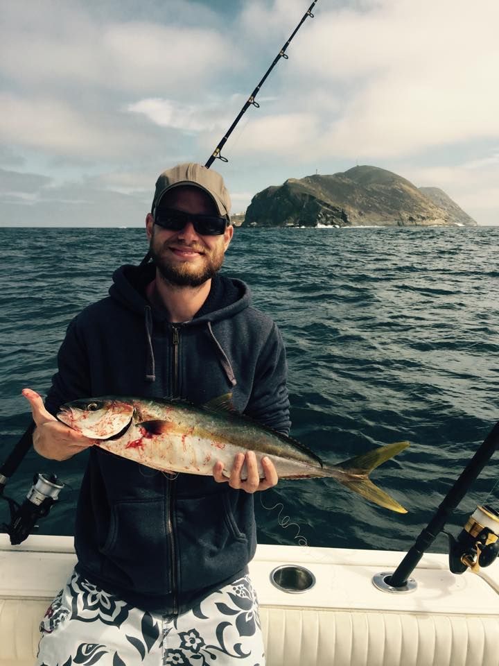 A person wearing sunglasses and a hoodie holds a caught fish while on a boat with a large coastal rock in the background.