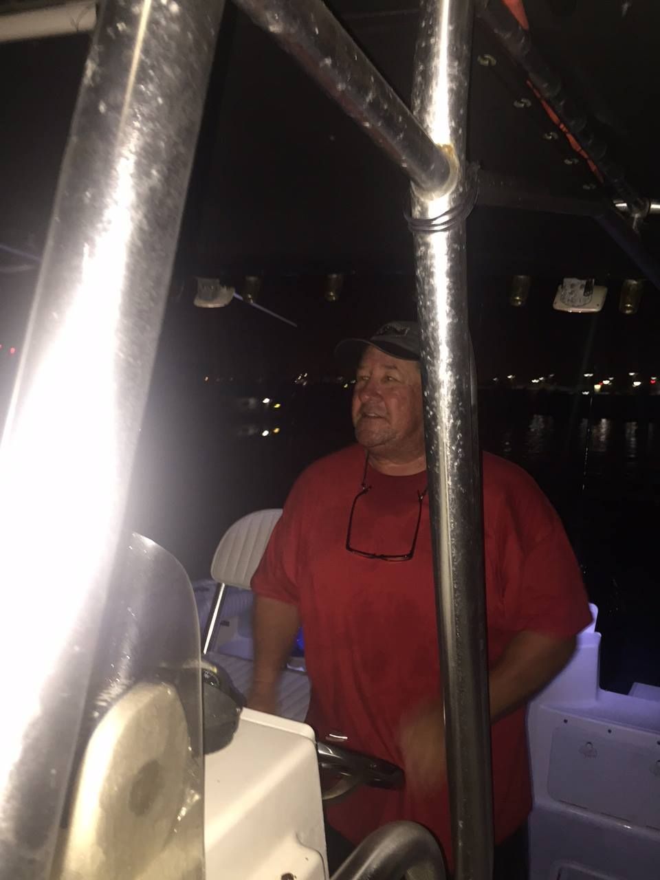 A person wearing a red shirt and cap at the helm of a boat at night, lit by interior lights.