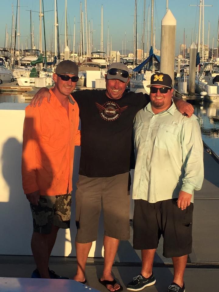 Three people in casual wear stand arm-in-arm on a marina dock, with many sailboats moored in the background.