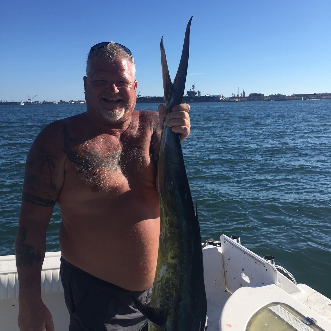 A shirtless man smiling while holding a large, freshly caught fish on a boat, with a harbor and large ship in the distance.