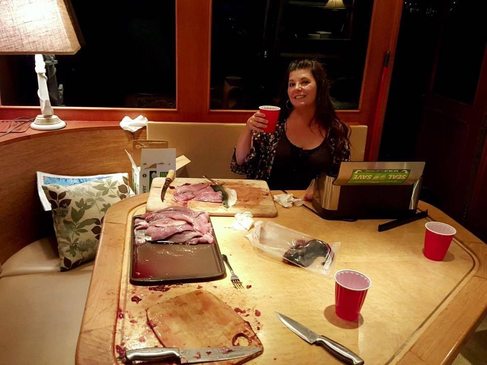 A person sits at a table with sliced meat, cutting boards, knives, and red plastic cups in a softly lit, wood-paneled room.