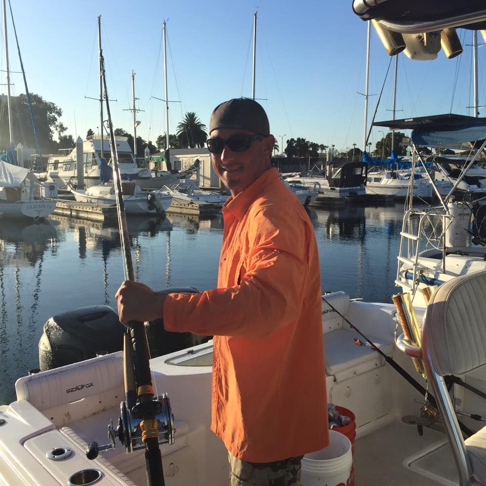 A person in a bright orange long-sleeved shirt and sunglasses holds a fishing rod while standing on a boat in a marina.