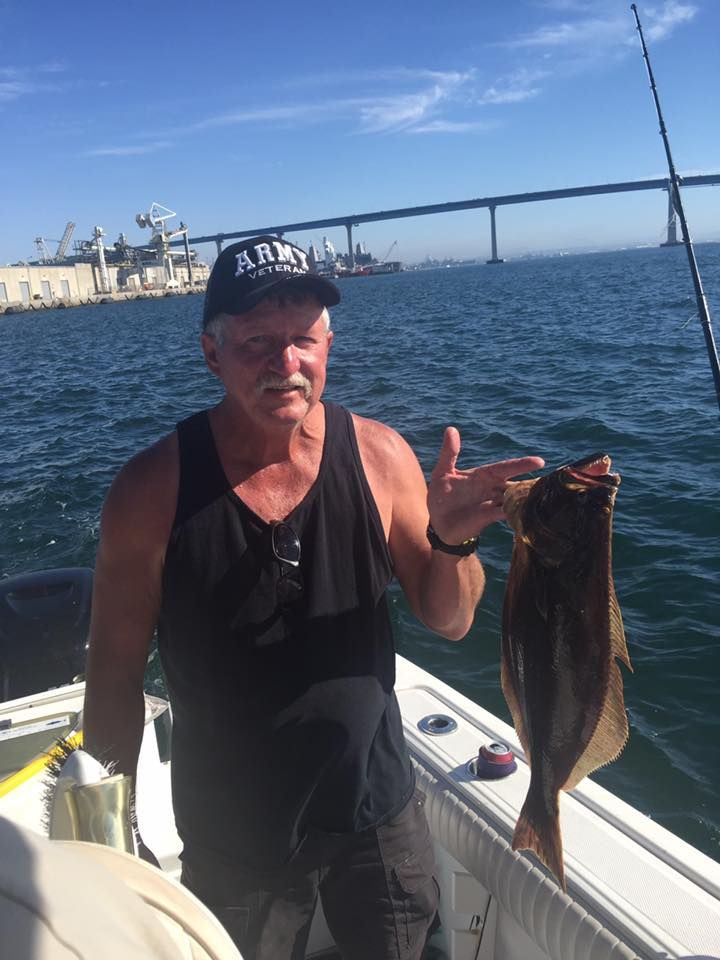 Man on boat holding fish, pointing toward it, with bridge and harbor in the background.